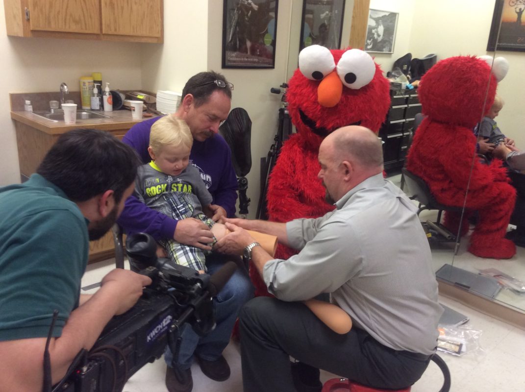 Steve Peeples at Peeples’ Orthotics and Prosthetics with Elmo (Adam Martinez from Shriners),  and grandpa Chris “Papa” Horning of Park City help Landon “Bear” Lacy in fitting his “legs.” Pic 3